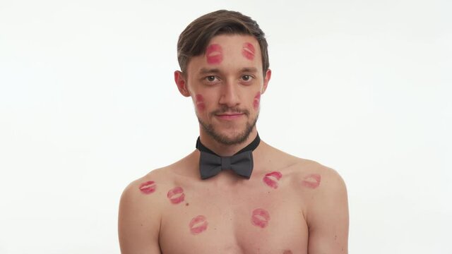 Close up portrait of one sexy model man wink twice. Half naked hot male with lipstick kiss marks on body and cheek wear grey bow tie, stand on white wall background. Happy Valentine’s day concept.