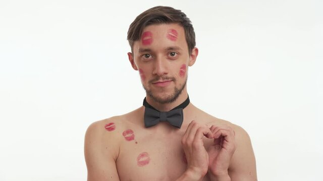 One young attractive European brunette man 20s with beard, red kiss imprints or marks on body and face wear grey bow tie makes a heart gesture with hands isolated on white wall background close up. 