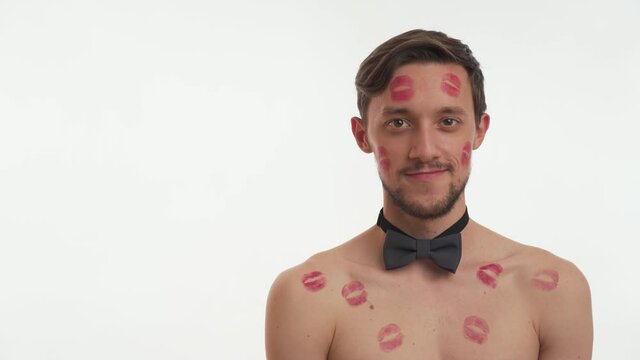 Attractive sexy European half naked brunette man 20s having beard and mustache, body covered with lipstick prints. Young guy smile, flirt, wink on white wall background. Romantic concept. Copy space.