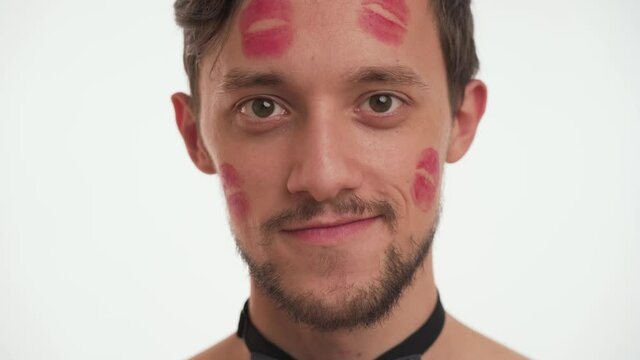 Closeup portrait of young handsome Caucasian brunette man with red kiss marks on face with brown beard and mustache look at camera, flirt, wink isolated on white wall background. Women’s day concept.