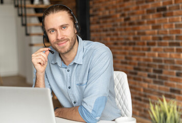 Millennial young male salesman in a headset is holding the microphone, hipster customer service representative posing against the brick wall, sitting in front of a laptop and looking at the camera
