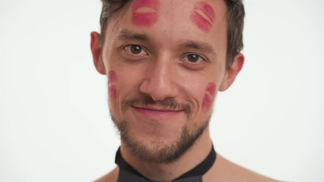 Cute young half naked man 30s wear black bow tie smile, flirt, make his eyes isolated on white background closeup. One handsome European male model face covered with red lipstick prints from girls.