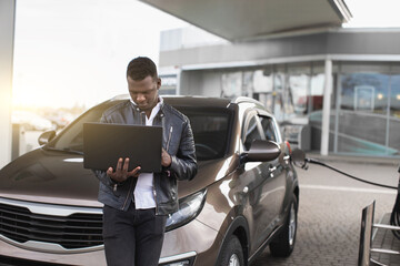 Young confient African American businessman, standing in front of his car and working on laptop pc,...