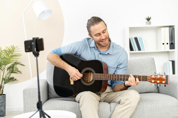 Handsome attractive bearded hipster guy making video blog about musical instruments, sitting on the couch, playing the guitar, and streaming tutorial online, using smartphone and tripod for recording