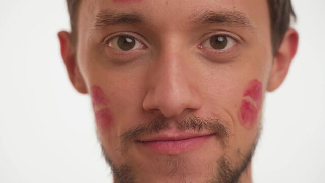 Gorgeous man with light in brown eyes, moustache, red kiss marks on face looking straight, smiling on white wall background close up. Portrait one handsome male in love with stains of lip gloss stick.