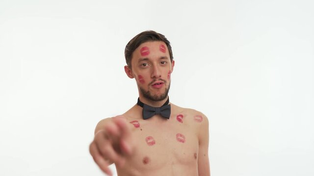 Blow kiss. Close up portrait of lovely brunette man with beard, stained red lipstick marks on body, face look at camera isolated on white background. Male stare, wear black bow tie. Romantic concept.