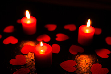 On a black textured background are scattered red hearts and stands three burning red candles, with blurry background, used as a background or texture, soft focus