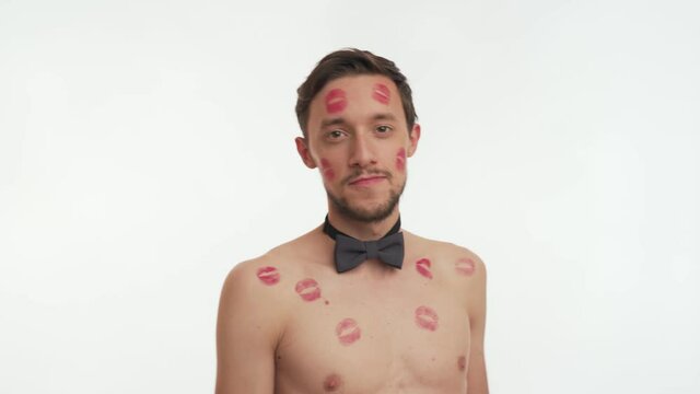 Young pleased European man with red lipstick kiss marks or stains on body get from girl wear dark bow tie, come closer on white background, smile, wink, leaves shot. Close up portrait of sensual male.