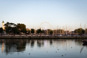 Sunset over harbor village with lighthouse and Ferris wheel in background.