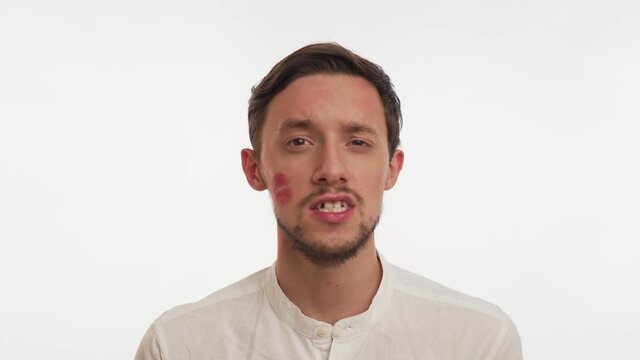 Caucasian brunette bearded man with red lips kiss mark, imprint on cheek wear shirt, show broken teeth isolated on white wall background. Close up portrait of confident lover business guy. Copy space.