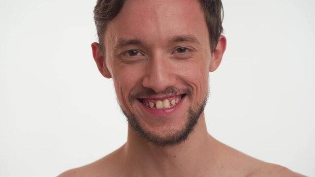 Close-up portrait Caucasian happy bearded half naked man with brown eyes, short hair, crooked broken teeth. Pleased brunette guy smiling isolated on white background. One male look at camera, laughs.