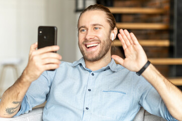 Smiling bearded hipster man sitting on the couch in the living room, video-calling, talking to friend, family, colleague, waving at the mobile phone, using earphones, staying connected during pandemic