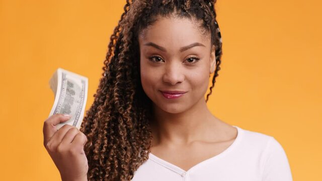 Mega profit. Confident young african american woman playing with stack of dollar banknotes, orange studio background