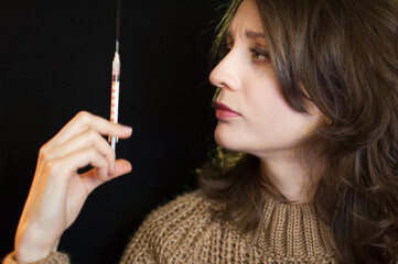 Obraz premium Vaccine concept. Female portrait of caucasian girl holding a syringe and looking at it on black background in studio.