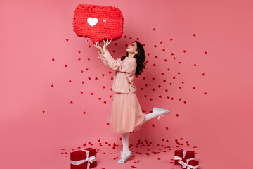 Inspired woman in skirt dancing with heart icon. Studio shot of carefree curly girl with presents, valentine's day concept.
