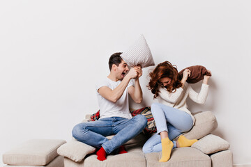 Excited young people enjoying pillow fight. Indoor shot of european couple fooling around at home.