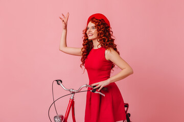Woman waving greeting while sitting on bicycle on pink background. Photo of lady in red beret and dress