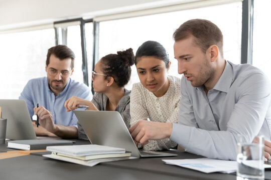 Team members discussing project at shared laptop in office. Millennial business colleagues working at computer together. Indian female coach training intern. Internship, teamwork concept