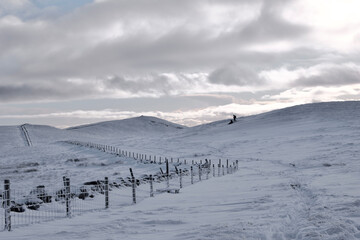 Fototapeta na wymiar snowy hilly landscape with a fence, stone wall and silhouettes of two hill walkers in the distance
