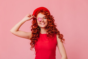 Lady in red top and hat posing with big candy. Curly redhead woman posing over pink background