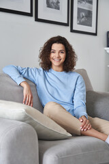 Smiling young pretty hispanic woman looking at camera sitting on comfortable sofa at home. Happy cheerful casual teen girl, female model posing indoors, relaxing on couch in modern apartment, portrait