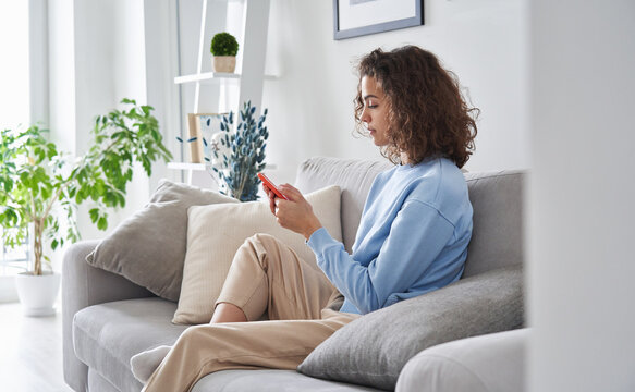 Hispanic Teen Girl Holding Cell Phone Sitting On Sofa At Home. Young Latin Woman Using Smartphone Tech Mobile Apps Online Services On Cellphone Device Relaxing In Apartment Living Room.