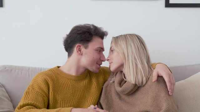 Happy affectionate young couple bonding, kissing, talking, cuddling, holding hands at home on sofa, enjoying hugging, chatting and relaxing together sitting on couch in cozy apartment living room.
