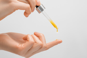 A pipette of orange-colored serum drips on the finger of a woman's hand, with a light background, a droplet with a glare in close-up.