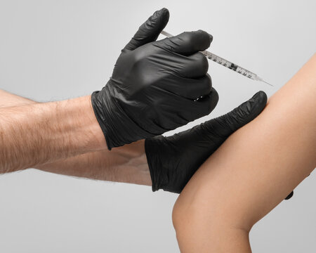 A male doctor wearing latex black gloves gives the vaccination to the patient in the shoulder, using a 1ml disposable syringe, close up.