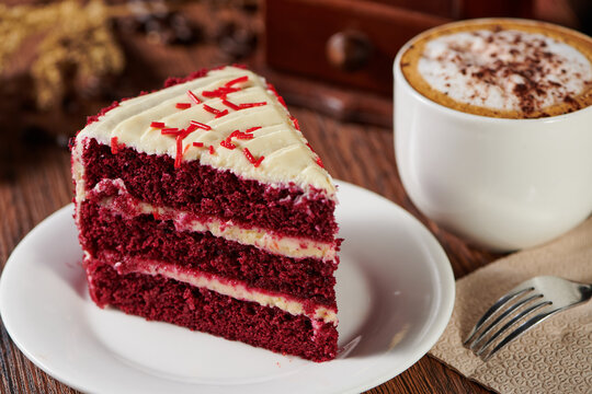 Slice of red velvet cake on a plate with a cappuccino on a wooden table