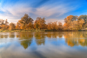 Beautiful violin shaped pier at autumn sunset over the river with reflected orange and green trees, clouds and blue sky
