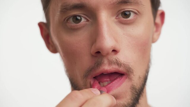 One young Caucasian attractive brunette man with brown eyes, beard holds pink lipstick clumsily paints own lips on white background close up. Portrait of gay looking directly at camera, use cosmetic.