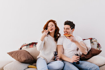 Young people sitting on couch and holding slice of pizza. Funny friends spending time at home.