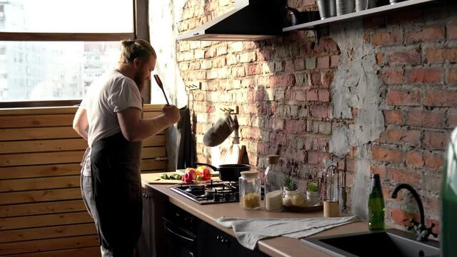 Happy male chef enjoying time at own kitchen dancing and singing using spatule as microphone, Caucasian man in apron preparing dinner dishes during daily routine in home apartment