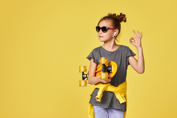 Stylish cool little child girl in sunglasses with yellow skateboard showing Ok gesture on yellow background. copy space