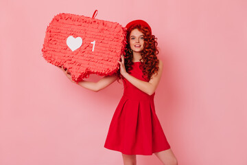 Fiery red curly lady in hat and dress is looking at camera and holding like sign on isolated background