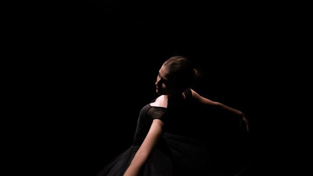 Orbital shot of a ballerina in the image of a black swan gracefully moving on a black studio background with backlight. Close up. Slow motion.