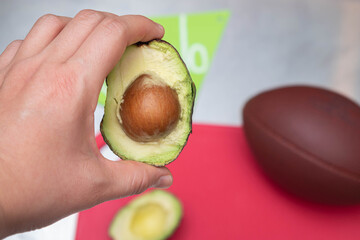 Woman hand holding an avocado cut in the middle. American football ball, red cutting board and banner on metal table - Powered by Adobe
