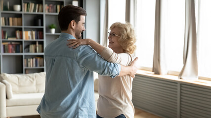 Fototapeta premium Smiling young caucasian man dancing with happy older middle aged beautiful mother in living room, enjoying weekend family hobby activity at home, grown children and retired parents pastime concept.