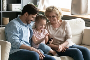 Happy young father and middle aged mature grandmother watching joyful little kid girl playing games on smartphone, enjoying using mobile applications together, sitting on cozy couch in living room.