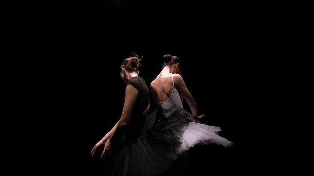Camera rotates around two flexible ballerinas in black and white tutus move their hands dramatically against a backlit black studio background. Orbital shot. Close up. Slow motion.