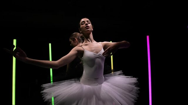Camera circular motion around young ballerinas dancing on a black studio background with bright neon lights. Elegant movements of a black and white swans in slow motion. Orbital shot close up.