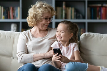 Happy bonding older mature grandmother sitting on comfortable couch with little preschool granddaughter, enjoying using smartphone, having fun playing mobile games together at home, modern tech usage.