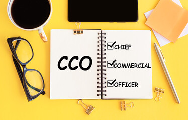 CCO - Chief Commercial officer. Text on notepad and office accessories on yellow desk.