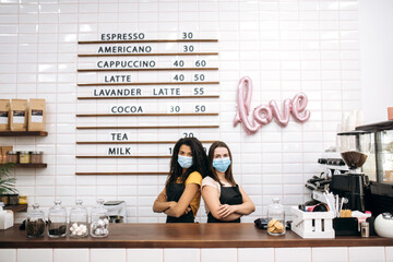 Two female waitress a coffee shop or restaurant, African American and Caucasian, stands behind the bar counter wearing medical masks, backs to each other with arms crossed and looking at the camera