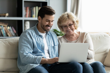Happy young man teaching smiling older retired mother using computer software applications at home. Joyful different generations family looking at laptop scree, enjoying web surfing or shopping online