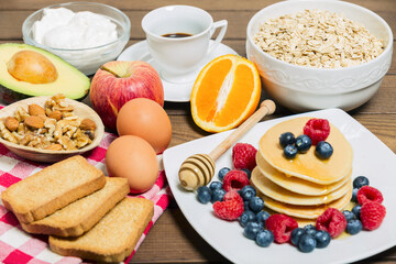Healthy breakfast background with coffee, pancakes, fresh berries, eggs, avocado, greek yogurt, oats, rusks and orange