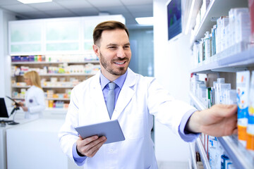 Naklejka na ściany i meble Handsome male pharmacist in white coat working in pharmacy store or drugstore. Checking medicines on his tablet computer. Healthcare and apothecary.