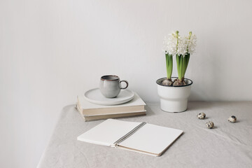 Easter spring still life. Cup of coffee, books and open diary mockup on linen table cloth. White hyacinth in flower pot. olive branches with quail eggs. Home office concept. Scandinavian interior.
