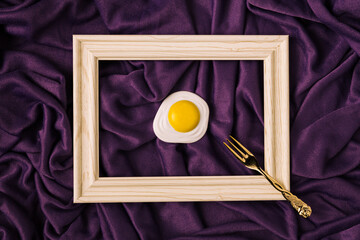 Abstract concept of backed egg with gold fork in the wood frame against elegant luxury purple satin or silk background in waves. 